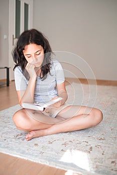 Pretty young girl reading paper book while sitting on carpet at home