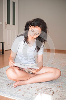 Pretty young girl reading paper book while sitting on carpet at home