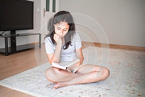 Pretty young girl reading paper book while sitting on carpet at home