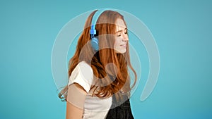 Pretty young girl with red hair listening to music, smiling, dancing in blue headphones in studio against plain