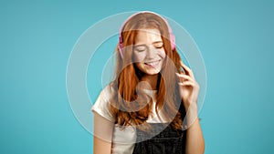 Pretty young girl with red hair listening to music, smiling, dancing in pink headphones in studio against blue