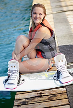 A pretty young girl sitting on a jetty next to a wakeboard