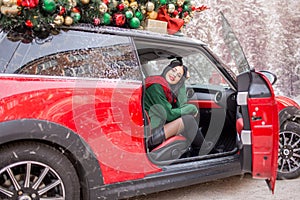 Pretty young girl is sitting in red car with decorated xmas tree on the roof.