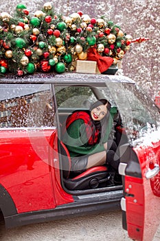 Pretty young girl is sitting in red car with decorated xmas tree on the roof.