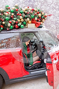 Pretty young girl is sitting in red car with decorated xmas tree on the roof.