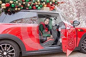 Pretty young girl is sitting in red car with decorated xmas tree on the roof.