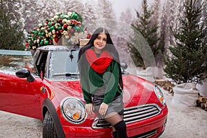 Pretty young girl is sitting on red car with decorated xmas tree on the roof, holiday concept.