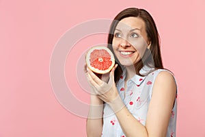 Pretty young girl in summer clothes looking up holding in hand half of fresh ripe grapefruit isolated on pink pastel