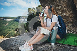 Pretty young girl traveling sitting in mountains drinking tea from camping thermos enjoying trip, tourist student is relaxing