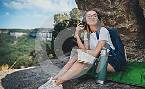 Pretty young girl traveling sitting in mountains planning route on map and drinking tea from camping thermos enjoying the trip