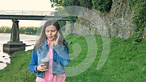 Pretty young girl walks along the waterfront talking on smartphone and drinking coffee. Bridge and vintage wall of wild