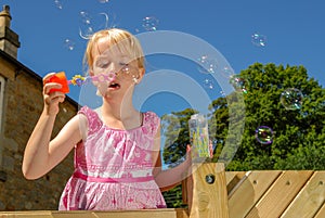 A pretty, young girl wearing a pink dress, blows bubbles from the top of her climbing frame, on a sunny summers day with a deep