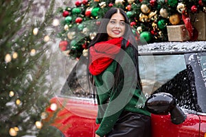 Pretty young girl is wearing red scarf sitting on red car with decorated xmas tree on the roof, holiday concept.