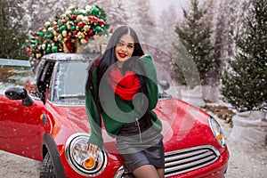 Pretty young girl is wearing red scarf sitting on red car with decorated xmas tree on the roof, holiday concept.