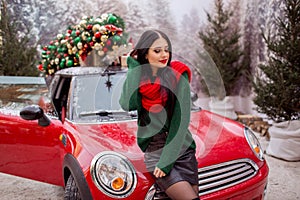 Pretty young girl is wearing red scarf sitting on red car with decorated xmas tree on the roof, holiday concept.