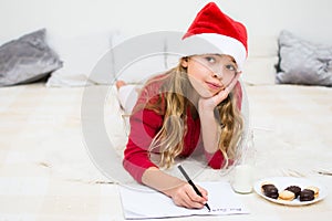 Pretty young girl writing letter for Santa Claus. Child in red santa hat lying on bed  and fhinking and writing christmas letter.