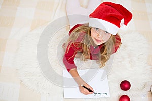 Pretty young girl writing letter for Santa Claus. Child in red santa hat lying on bed and writing christmas letter. Christmas