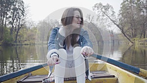 Pretty young glamour woman in white pants, jeans jacket and sunglasses paddles on the yellow boat on the river, looking