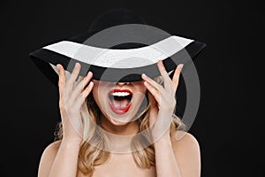 Pretty young happy blonde woman with bright makeup red lips posing isolated over black wall background wearing hat