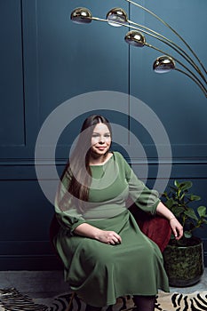 Pretty young lady in a evening dress sitting in a chair on dark blue background. Plus size caucasian female model posing