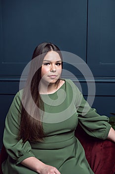 Pretty young lady in a evening dress sitting in a chair on dark blue background. Plus size caucasian female model posing