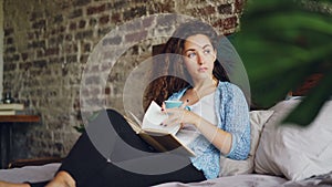 Pretty young lady is reading book in bed holding cup of tea and resting on pillows enjoying day at home. Beautiful loft