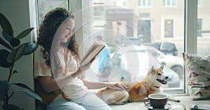 Pretty young lady reading novel in cafe on window sill and stroking pet dog