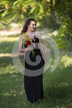 Pretty young lady stands under a tree holding a bouquet of wild flowers