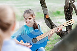 Pretty young little girl playing acoustic guitar for her friend in outdoors spring park