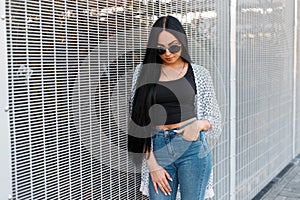 Pretty young modern hipster woman model with long hair in an elegant jacket in round sunglasses in a black T-shirt in jeans