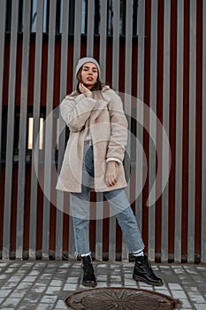 Pretty young modern woman in a fashionable cozy faux fur coat milk color in stylish blue jeans in a vintage knitted hat in trendy