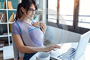 Pretty young mother with her baby in sling working with laptop at home