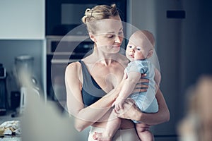 Pretty young mother holding her newborn baby boy standing near kitchen window at home