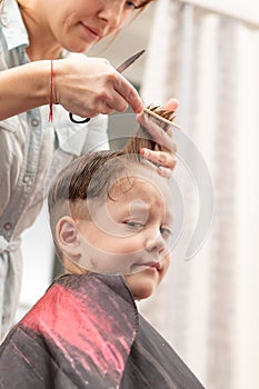 Pretty young mother in a light blue dress with a comb and scissors in her hands cuts her son`s hair at home. selective focus