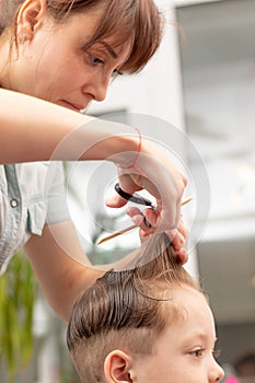 Pretty young mother in a light blue dress with a comb and scissors in her hands cuts her son`s hair at home. selective focus
