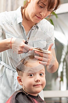pretty young mother in a light blue dress with a comb and scissors in her hands cuts her son`s hair at home. selective focus