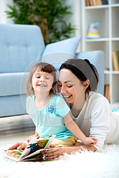 Pretty young mother reading a book to her daughter sitting on the carpet on the floor in the room. Reading with children