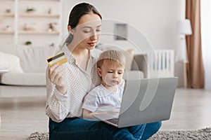 Pretty young mother with toddler baby using laptop and credit card, shopping online, ordering toys or clothes from home