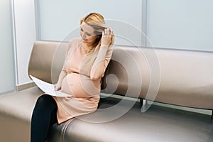 Pretty young pregnant woman is sitting in hallway of hospital waiting to see gynecologist.