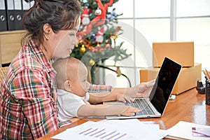 Pretty young single mom working at home on a laptop computer while holding her baby girl sitting on her lap enjoying playing