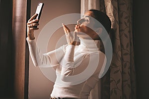 Pretty young smiled Caucasian woman in white turtleneck smiling and videochatting on mobile phone in home.Girl talking