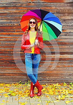 Pretty young smiling woman with colorful umbrella wearing a red leather jacket and rubber boots in autumn over wooden background