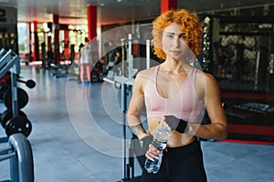 Pretty young sportswoman relaxing after training and drinking water in gym