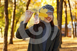 Pretty young stylish happy woman with a smile in a fashionable coat with a vintage hat with a yellow autumn leaf covers her face