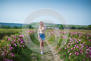 Pretty Young tender woman walking in the tea roses field. Blond lady wearing jeans and retro hat enjoy summer day