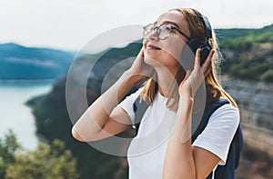 Pretty young tourist woman with backpack and  hipster glasses closes her eyes listens favorite music with headphones enjoy