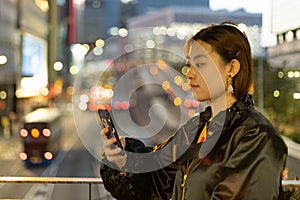 Pretty young university student girl read mobile phone in Central, Hong Kong with night view of blurred colorful city lights in