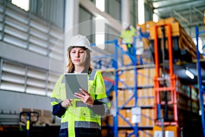 Pretty young warehouse worker woman hold tablet and look at tablet and her co-worker work on the background during carry product