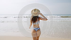 Pretty young woman applying sunscreen on face and running to sea. Attractive fitted girl in bikini and straw hat having