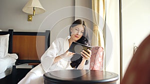 Pretty young woman in bathrobe chatting on tablet computer sitting on chair in hotel room